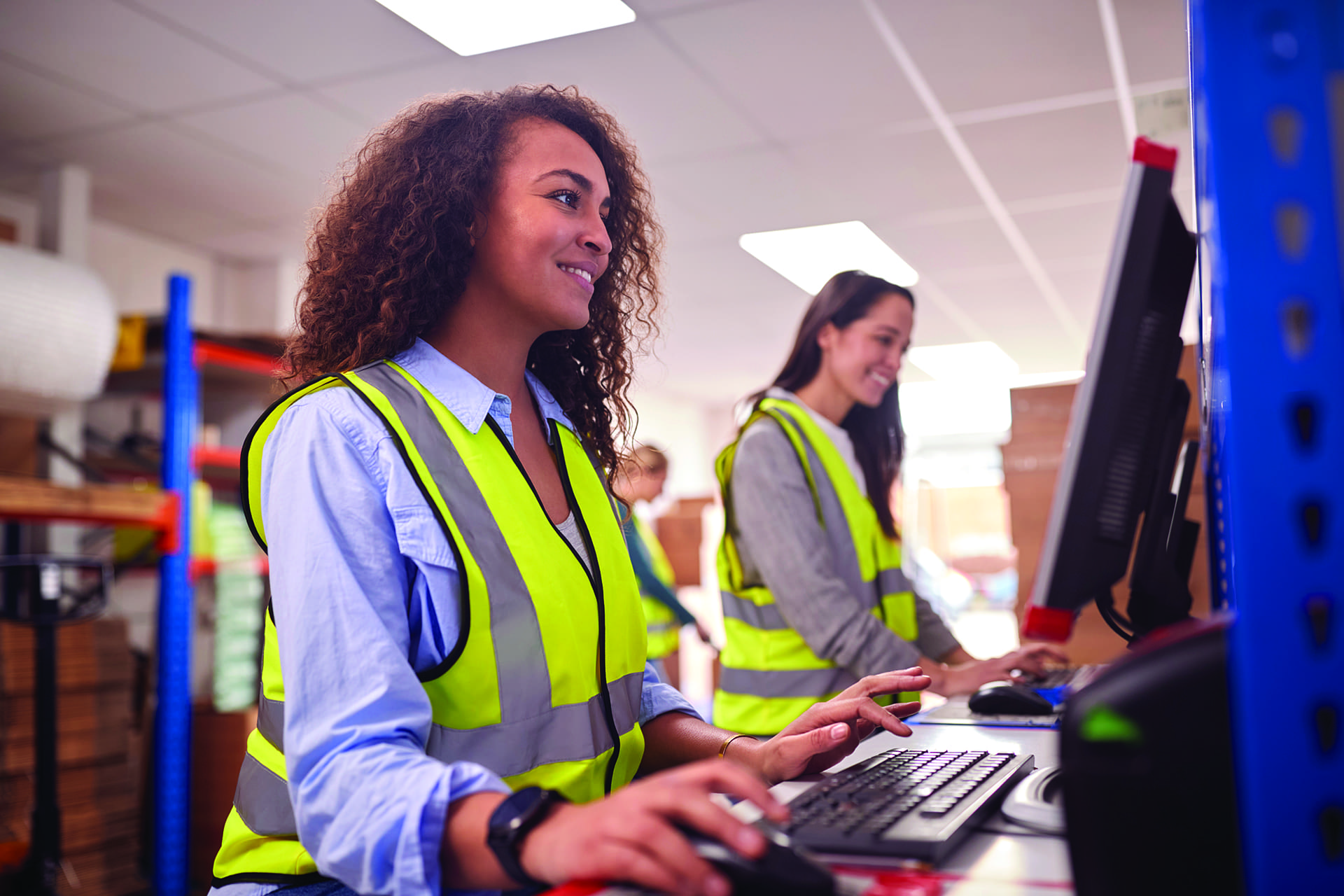 Female Customer Service Staff In Busy Modern Warehouse Working On Computer Terminals